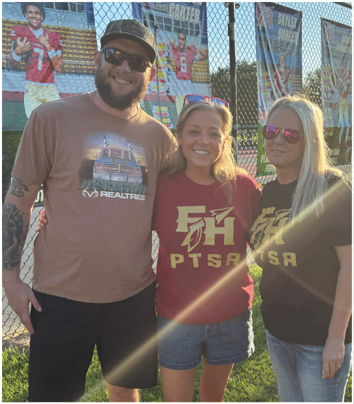 Two adults wearing matching PTSA t-shirts stand outdoors near a chain-link fence with sunlight creating a lens flare across the image. Description generated by AI