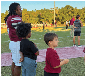 Two children stand side by side on a grassy field with one child wearing a black shirt and blue shorts and the other wearing a red shirt. Description generated by AI