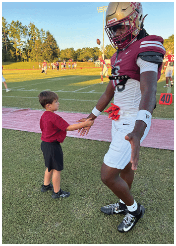 A child wearing a red shirt and black shorts stands on a sports field near a pink mat while a group of people in sports uniforms gather in the background. Description generated by AI