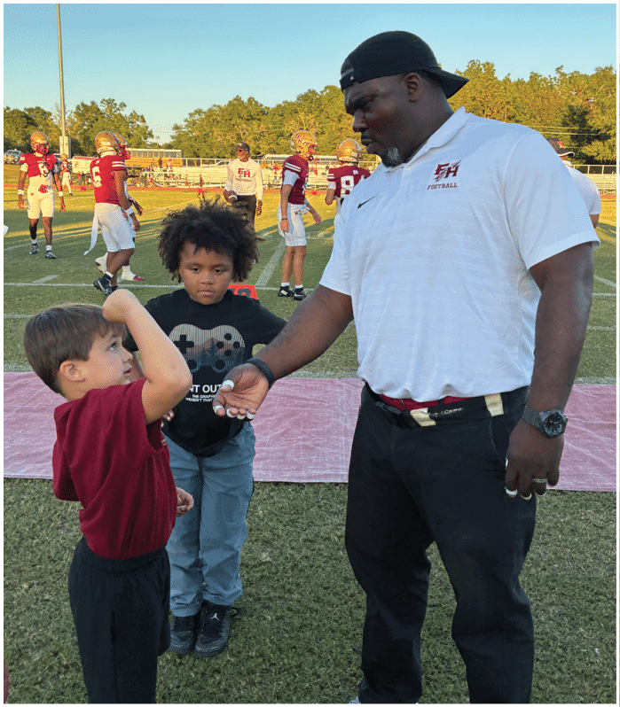 An adult wearing a white football polo and black cap is handing something to a child with curly hair standing on a sports field with other people in football uniforms in the background. Description generated by AI