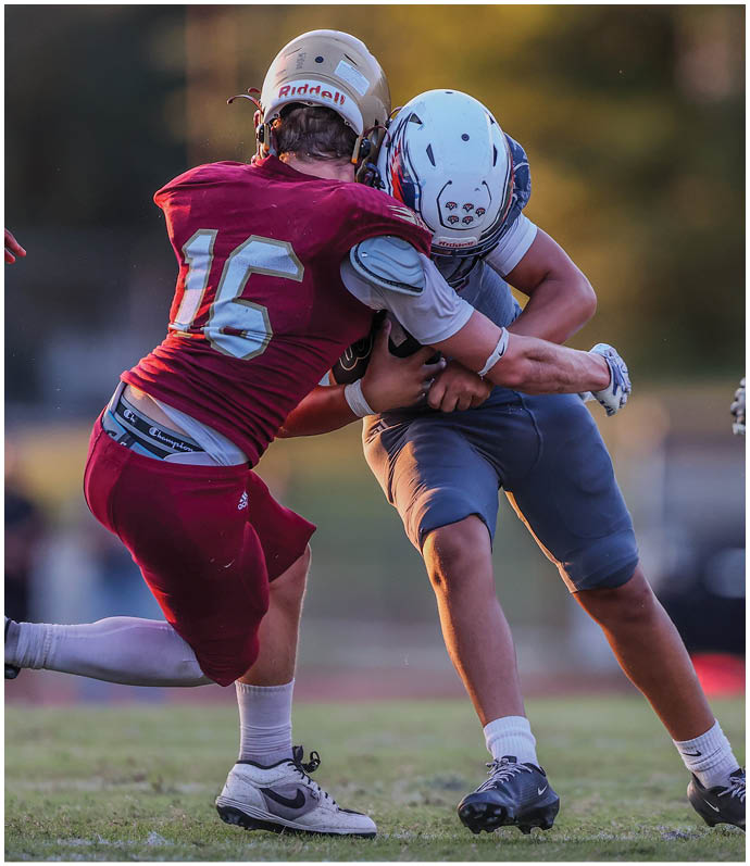 Two people in football uniforms are engaged in a tackle on a grassy field during a game. Description generated by AI