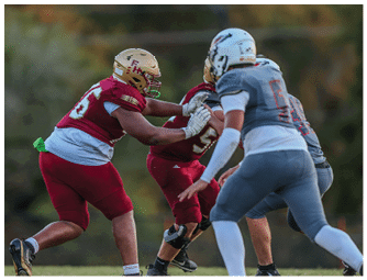 People wearing football uniforms are actively engaged in a play on a grassy field with trees in the background. Description generated by AI