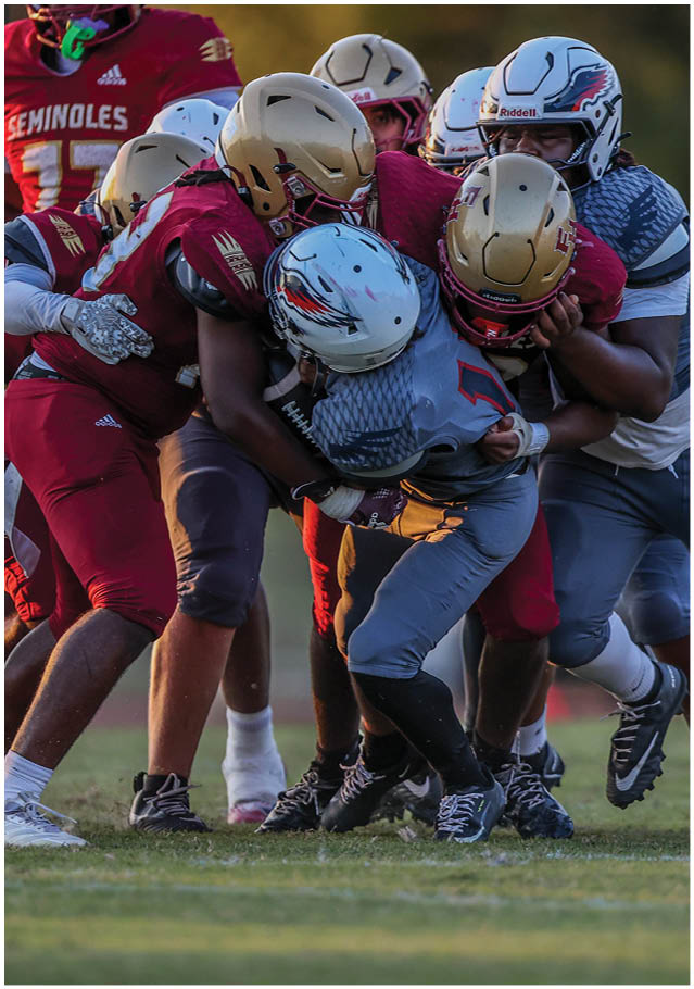 Several people wearing football helmets and uniforms are engaged in a close physical tackle on a grassy field during a game. Description generated by AI