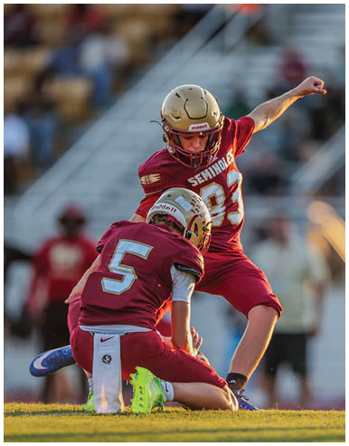 Two people in football uniforms with helmets are on a field, one kneeling and holding the ball while the other is preparing to kick it. Description generated by AI