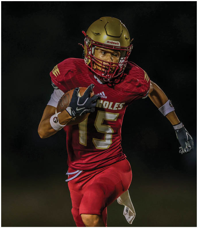 A person wearing a red football uniform with the number 15 and a gold helmet is running while holding a football. Description generated by AI