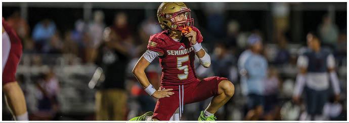 A person wearing a red football uniform with the number five and a gold helmet is kneeling on one knee on a field with blurred spectators in the background. Description generated by AI
