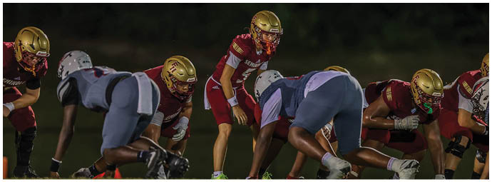 People in football uniforms are lined up on a field preparing for a play during a game at night. Description generated by AI