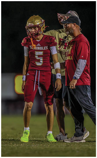 A person in a football uniform with the number five stands on a field while two adults in casual sportswear stand beside them, one placing a hand on their shoulder. Description generated by AI