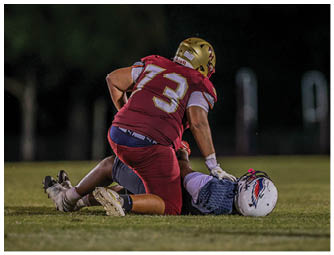 A person wearing a maroon sports uniform with the number 73 is kneeling on the ground while holding another person lying on the grass wearing a white helmet and a blue jersey during a nighttime outdoor sports event. Description generated by AI