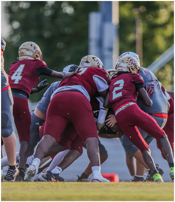 Several people wearing football helmets and uniforms are engaged in a close physical play on a grassy field during a game. Description generated by AI