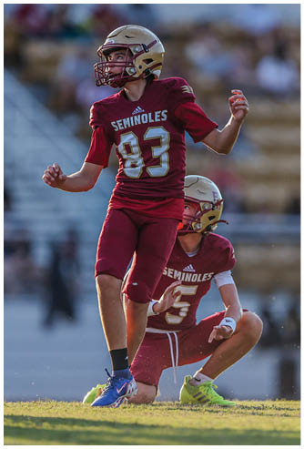 Two people wearing maroon football uniforms with “Seminoles" on the jerseys are engaged in a football play, with one person preparing to kick the ball and the other holding it on the ground. Description generated by AI