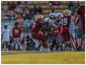 Two people in maroon and gold football uniforms tackle a person in a gray uniform holding a football on a field with blurred spectators in the background. Description generated by AI