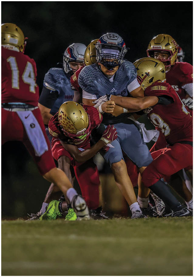 Several people in football uniforms are engaged in a tackle during a nighttime game on a grassy field. Description generated by AI