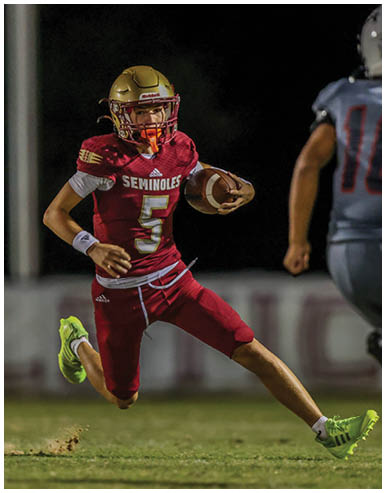 A person wearing a maroon football uniform with the word Seminoles and the number 5 is running on a grass field while holding a football. Description generated by AI