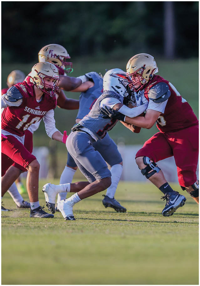 Two people in football uniforms and helmets are engaged in a physical tackle on a grassy field with other players in the background. Description generated by AI