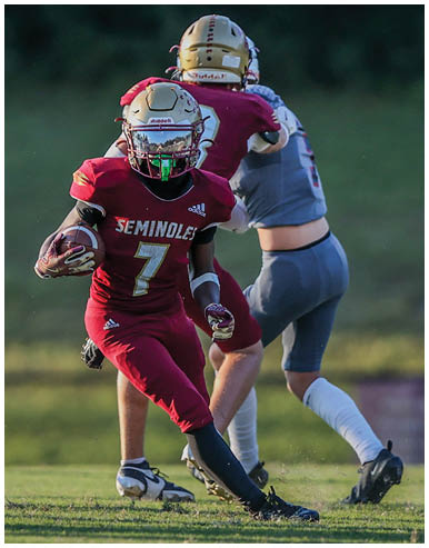 A person in a red football uniform with the number seven is running with the ball on a grassy field while another person in a gray uniform is behind them. Description generated by AI