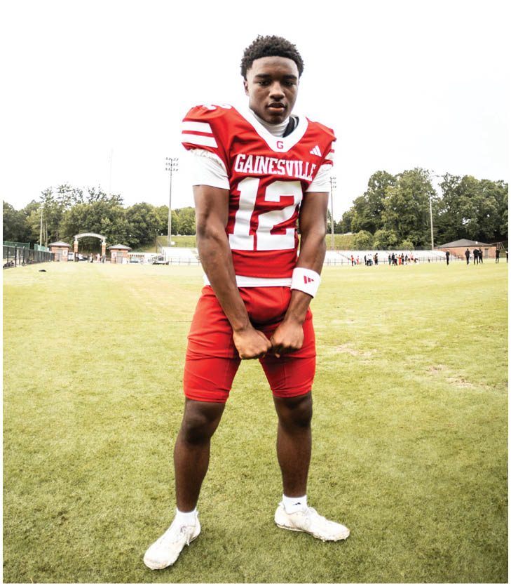 A person wearing a red and white football uniform with the number 12 stands on a grassy field with trees and people in the background. Description generated by AI