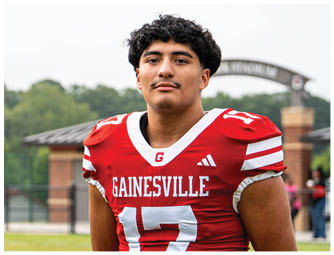 Two people wearing red Gainesville football uniforms stand on a grassy field with a sports facility in the background. Description generated by AI