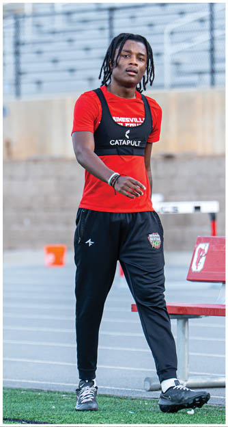 A person with braided hair wearing a red shirt and black athletic pants stands on a track field near a bench and hurdles. Description generated by AI