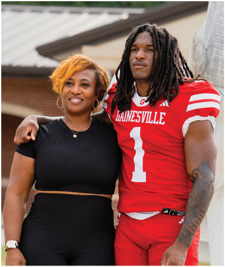 A person in a red football uniform with the word Gainesville and the number one stands with an arm around another person wearing a black outfit, both smiling outdoors. Description generated by AI