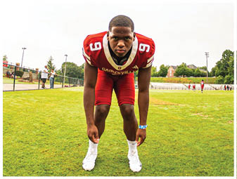 A person wearing a red football uniform with the number 9 is crouched in a ready position on a grassy field with a fence and trees in the background. Description generated by AI
