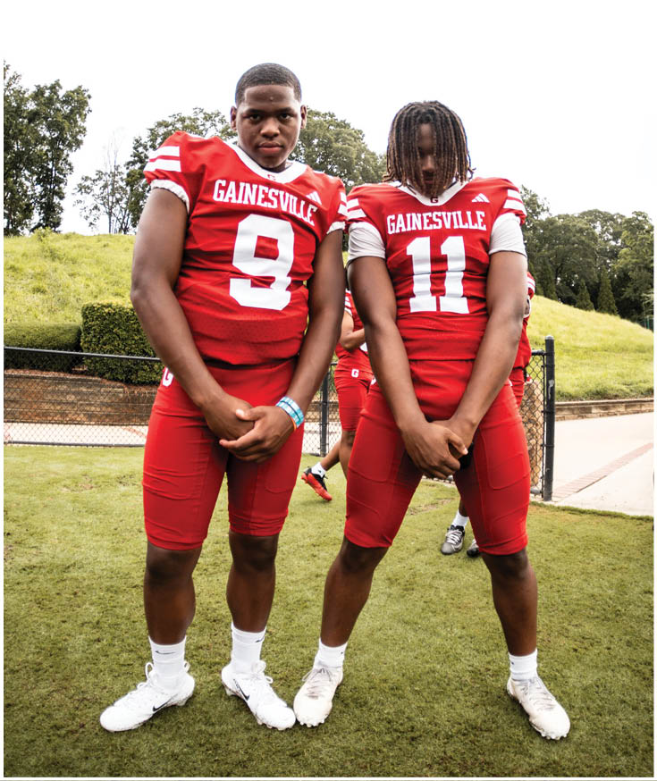 Two people wearing red Gainesville football uniforms stand on grass with their hands clasped in front of them while two others stand in the background near a fence. Description generated by AI