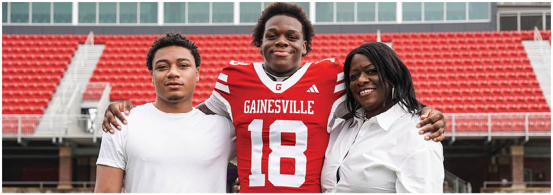 Three people stand closely together in a sports stadium with one person wearing a red Gainesville football jersey and the others in casual white shirts, all smiling. Description generated by AI