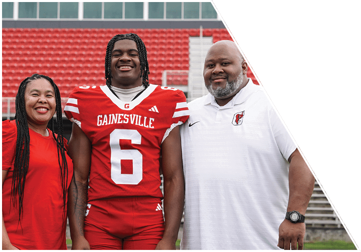 Three people stand together smiling on a sports field with one person wearing a red football uniform and the other two wearing casual clothing. Description generated by AI