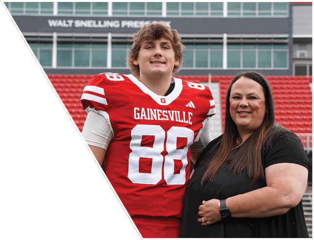 A person wearing a red Gainesville football jersey stands next to another person in a black dress on a sports field with red bleachers in the background. Description generated by AI