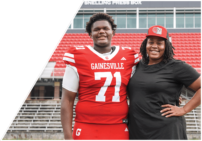 Two people stand side by side on a sports field, one wearing a red athletic uniform and the other wearing a black shirt and jeans. Description generated by AI