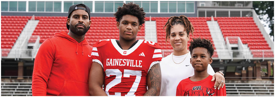 Four people stand together in a sports stadium with one person wearing a red football uniform and the others dressed casually. Description generated by AI