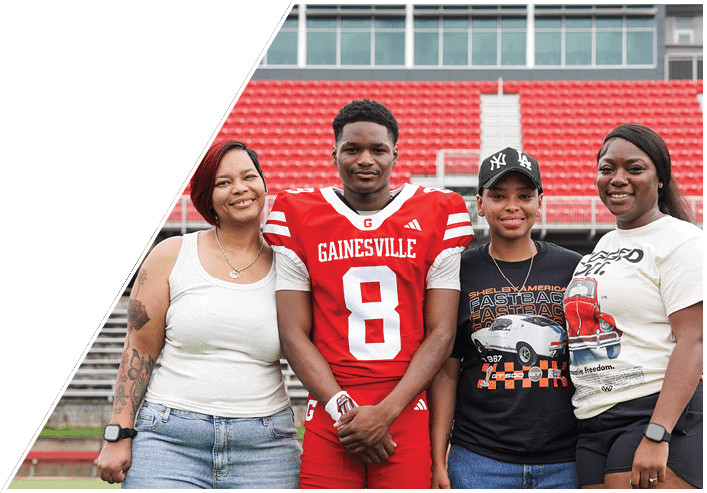 Four people stand together smiling on a sports field with red stadium seats in the background, one wearing a red football uniform and the others in casual clothing. Description generated by AI