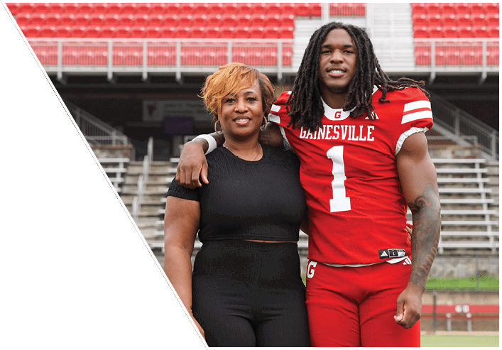 An adult wearing a red football uniform with the word “Gainesville" and the number one stands with an arm around another adult dressed in black in a stadium with empty red seats and bleachers. Description generated by AI