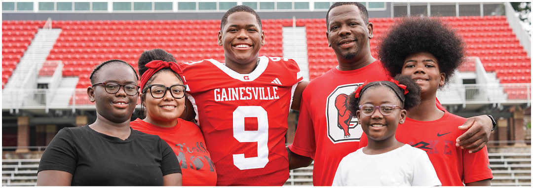 A group of six smiling people, including one wearing a red Gainesville sports jersey, stand closely together in a stadium with red seats in the background. Description generated by AI