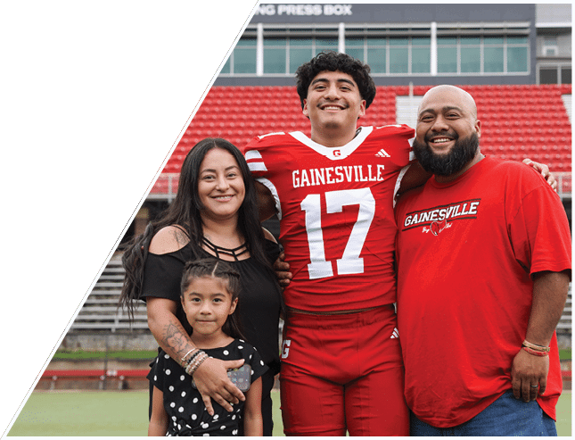 A group of four people, including one wearing a red football uniform with the number 17 and the word Gainesville, stand closely together smiling in a stadium with red seats. Description generated by AI