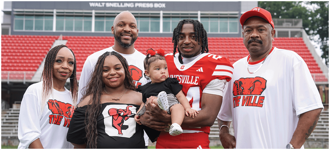 A group of six people, including one holding a child and one wearing a sports jersey, stand together on a sports field with stadium seating in the background. Description generated by AI