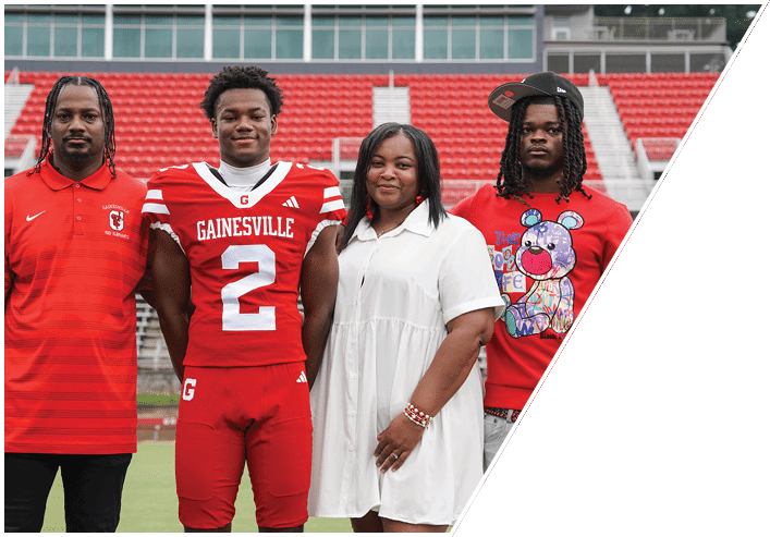 Four people stand together on a sports field with red bleachers in the background, including one person wearing a red football uniform with the word Gainesville and the number 2 on it. Description generated by AI