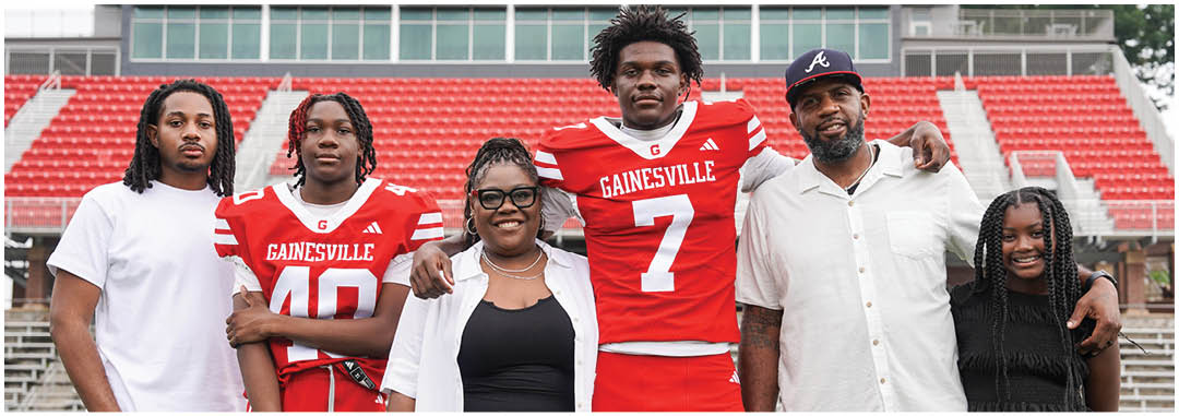 Five adults and one child stand together on a sports field with red bleachers in the background, two adults wearing red sports jerseys with “Gainesville" written on them. Description generated by AI