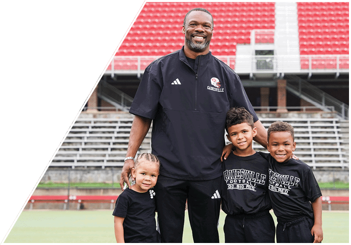 An adult wearing a sports jacket stands on a field smiling with three children dressed in matching sports shirts, with empty stadium seats in the background. Description generated by AI
