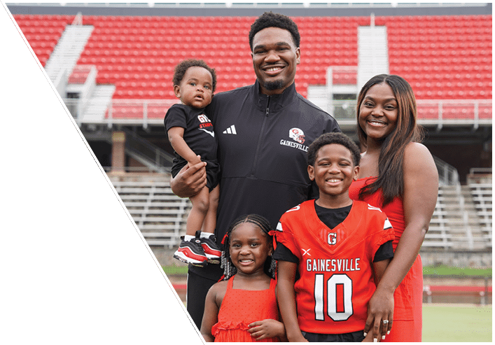 A group of five people, including two children wearing red Gainesville sports jerseys and three adults smiling together on a sports field with stadium seating in the background. Description generated by AI