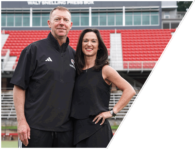 Two adults smiling and standing close together in front of stadium seating with a sign that reads “Walt Snelling Press Box" in the background. Description generated by AI
