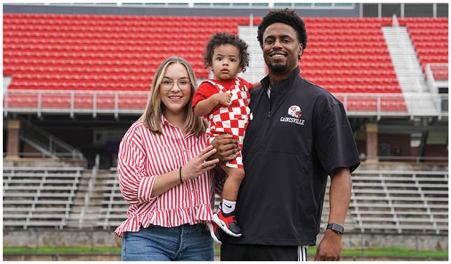 Two adults and a child stand in front of empty stadium seats with one adult holding the child who is wearing a red and white checkered outfit. Description generated by AI
