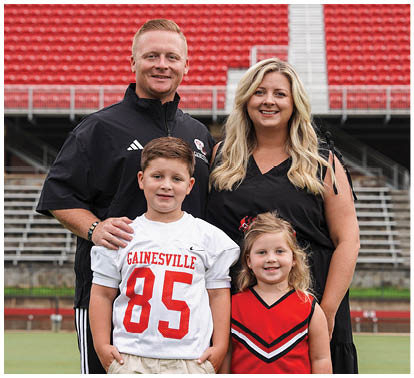 A group of four people, two adults and two children, stand smiling on a sports field with stadium seating in the background, with one child wearing a sports jersey and the other in a cheerleader outfit. Description generated by AI