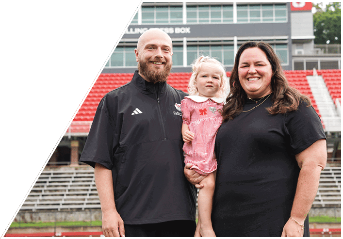 Three people, including one holding a child dressed in a pink outfit, stand smiling in front of stadium seating. Description generated by AI