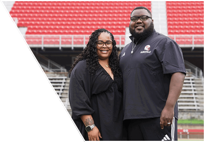 Two adults wearing glasses and casual clothing stand closely together smiling in an outdoor stadium with red and gray bleachers in the background. Description generated by AI