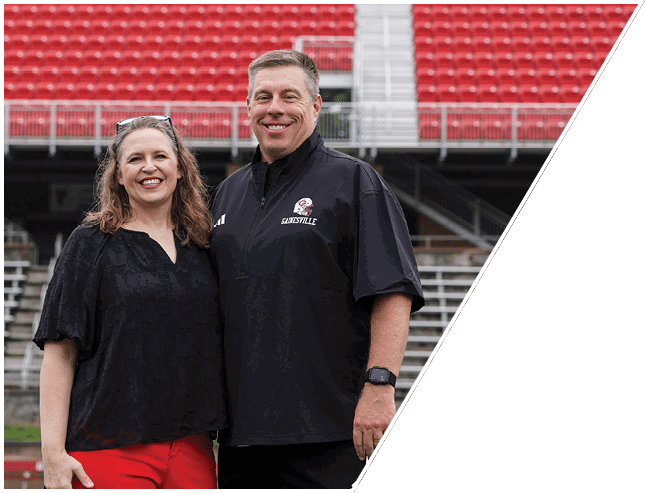 Two adults smiling and standing close together on a sports field with red stadium seats in the background. Description generated by AI