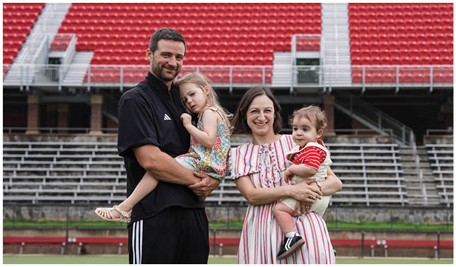 Two adults standing on a sports field holding a child each, with one adult wearing a black outfit and the other wearing a striped dress, both smiling. Description generated by AI