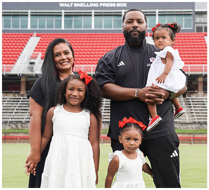 A group of five people, including three children wearing white dresses with red bows and two adults standing on a sports field with red stadium seats in the background. Description generated by AI