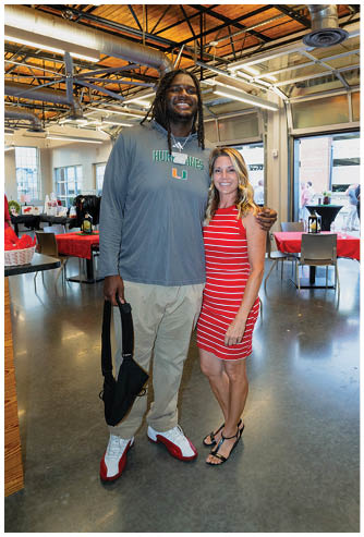 Two people stand smiling together in a spacious room with wooden ceilings and industrial lighting, one wearing a long-sleeve shirt and pants holding a bag, and the other wearing a red and white striped dress and sandals. Description generated by AI