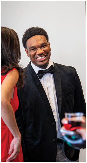 A person wearing a black velvet jacket and bow tie is smiling while standing next to two other people, one holding a glass of red wine. Description generated by AI